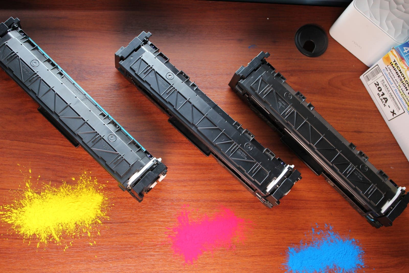 Tres cartuchos de tóner negros sobre una mesa de madera con tóner amarillo, magenta y cian derramado.